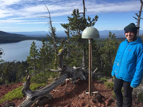 Maggie Holahan posing for the camera with a lake, forest, and mountains in the background